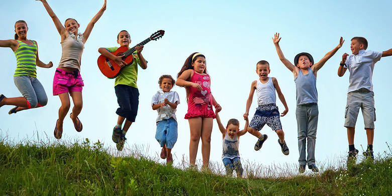 Kindergruppe die hüpfend in einer Wiese steht, mit blauen Himmel im Hintergrund - einer hat eine Gitarre in der Hand